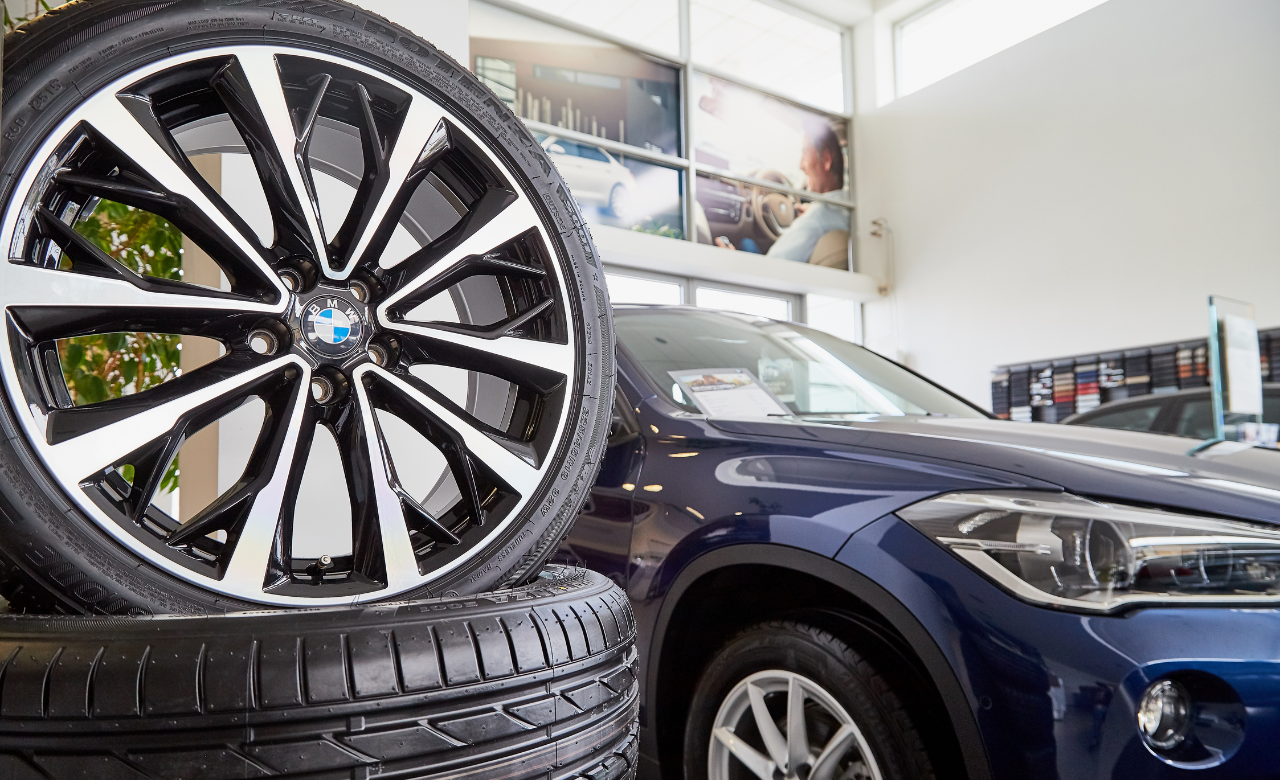 Close-up of a BMW wheel rim displayed on stacked tires in a bright showroom. A blue BMW SUV is partially visible in the background. Large windows provide natural light, and a blurred photo of a person driving is seen on the wall. Euro Mechanic | Bloor West Auto Repair Mechanics | Top Reviewed Auto Repair Shop in Mississauga | Visit bloorwestauto.ca