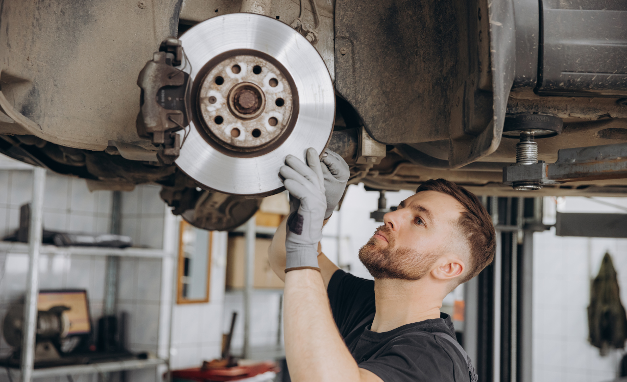 Mechanic in a workshop inspecting and adjusting a cars disc brake. The car is lifted on a hoist. The mechanic is wearing gloves and focused on the brake components. Various tools and equipment are visible in the background. Euro Mechanic | Bloor West Auto Repair Mechanics | Top Reviewed Auto Repair Shop in Mississauga | Visit bloorwestauto.ca