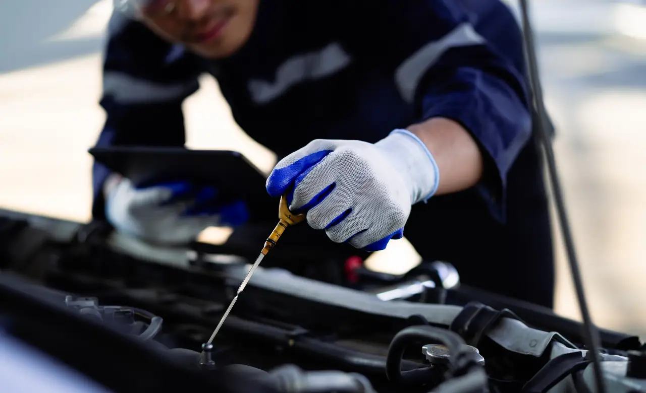 A mechanic in a dark blue uniform with reflective stripes and white gloves holds a dipstick over a car engine, checking the oil level. They are also holding a digital tablet, focusing on the task. Euro Mechanic | Bloor West Auto Repair Mechanics | Top Reviewed Auto Repair Shop in Mississauga | Visit bloorwestauto.ca