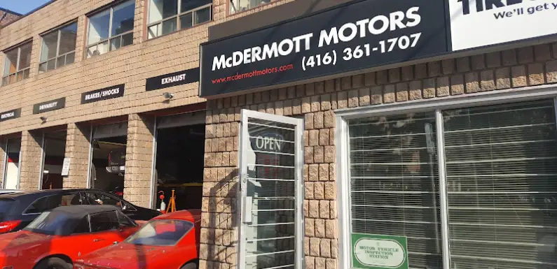 The exterior of McDermott Motors showcases a sign with their phone number. Garage doors labeled for brakes, shocks, and exhaust are open, revealing cars inside. A red car is parked outside under the OPEN sign on the glass door, marking it as a key location in Bloor West Autos bustling scene. Euro Mechanic | Bloor West Auto Repair Mechanics | Top Reviewed Auto Repair Shop in Mississauga | Visit bloorwestauto.ca
