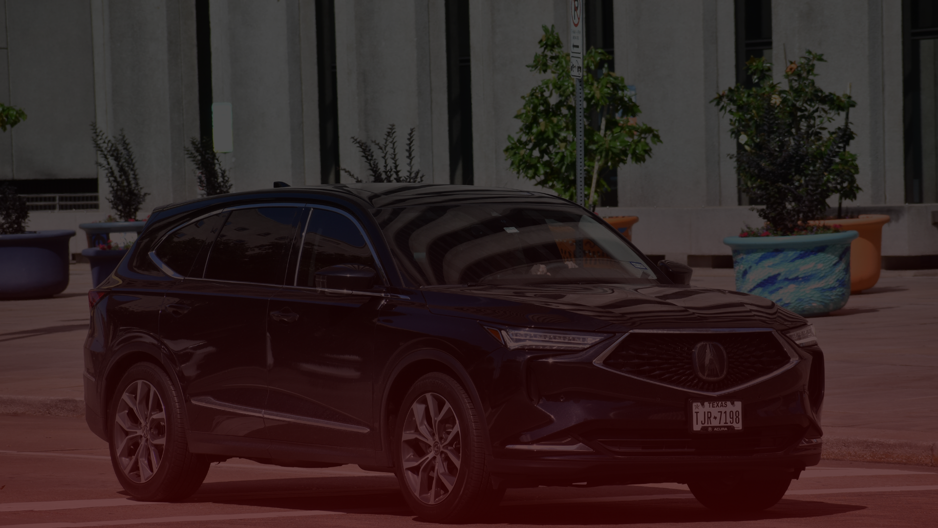 A dark-colored SUV is parked on a street in front of a modern building with large windows, hinting at the sleek destinations it frequents for Acura repair in Mississauga. Several colorful planters with small trees and plants add vibrancy near the building. The car has visible license plates. Euro Mechanic | Bloor West Auto Repair Mechanics | Top Reviewed Auto Repair Shop in Mississauga | Visit bloorwestauto.ca