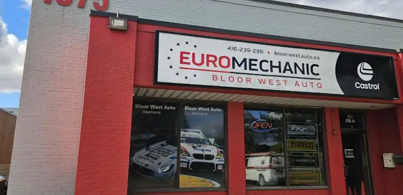 A red and white building marked Euromechanic Bloor West Auto stands proudly with its contact details displayed. Large windows showcase automotive posters and decals beneath a glowing neon Open sign, while the Castrol logo graces the right side, emphasizing its prime location. Euro Mechanic | Bloor West Auto Repair Mechanics | Top Reviewed Auto Repair Shop in Mississauga | Visit bloorwestauto.ca