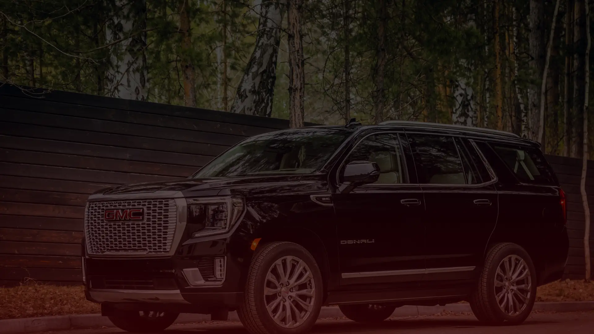 A black GMC Denali SUV, emblematic of precision found at a GM Mechanic in Mississauga, is parked in front of a tall black fence with trees in the background. The vehicle faces the camera, showcasing its grille and headlights, set against a serene wooded area. Euro Mechanic | Bloor West Auto Repair Mechanics | Top Reviewed Auto Repair Shop in Mississauga | Visit bloorwestauto.ca