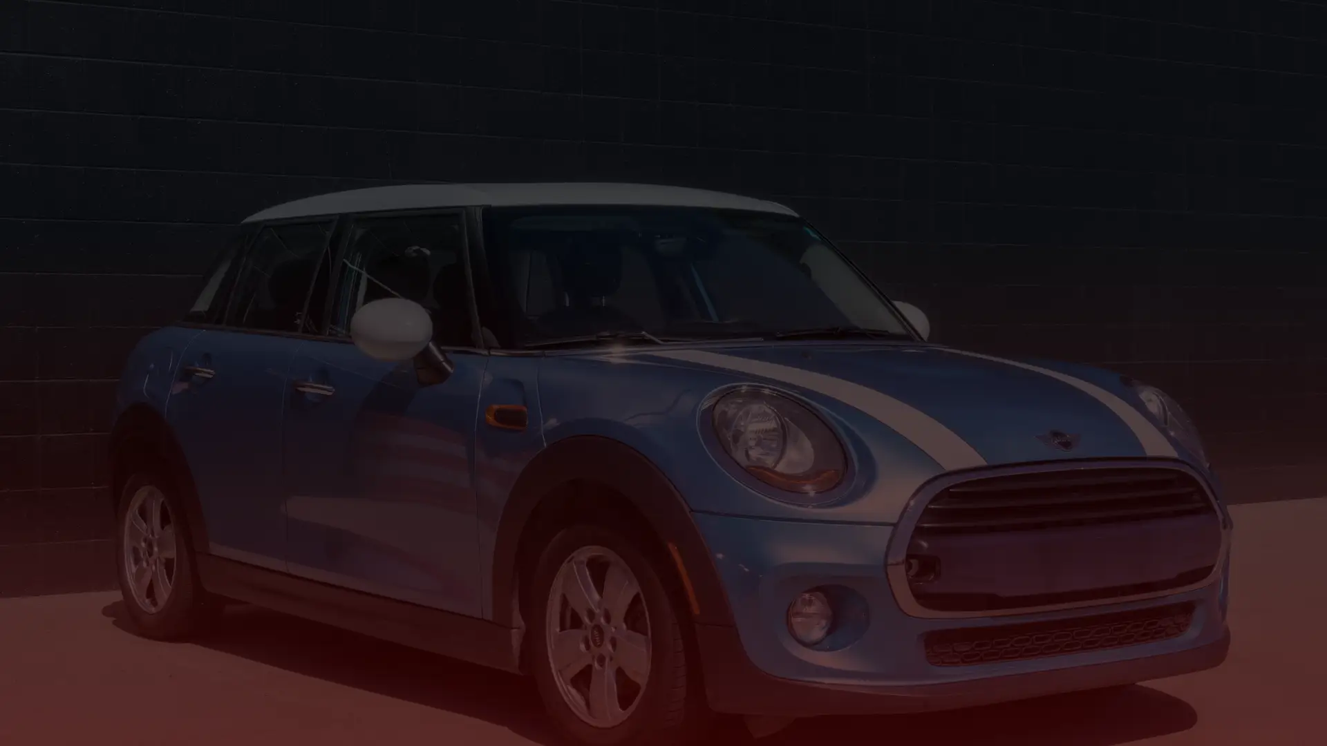 A blue Mini Cooper with white racing stripes is parked in front of a dark, textured wall, its design featuring a white roof and matching side mirrors. The sun casts shadows across its sleek body, awaiting a tune-up at Bloor West Auto’s expert Mini Cooper Repair service. Euro Mechanic | Bloor West Auto Repair Mechanics | Top Reviewed Auto Repair Shop in Mississauga | Visit bloorwestauto.ca